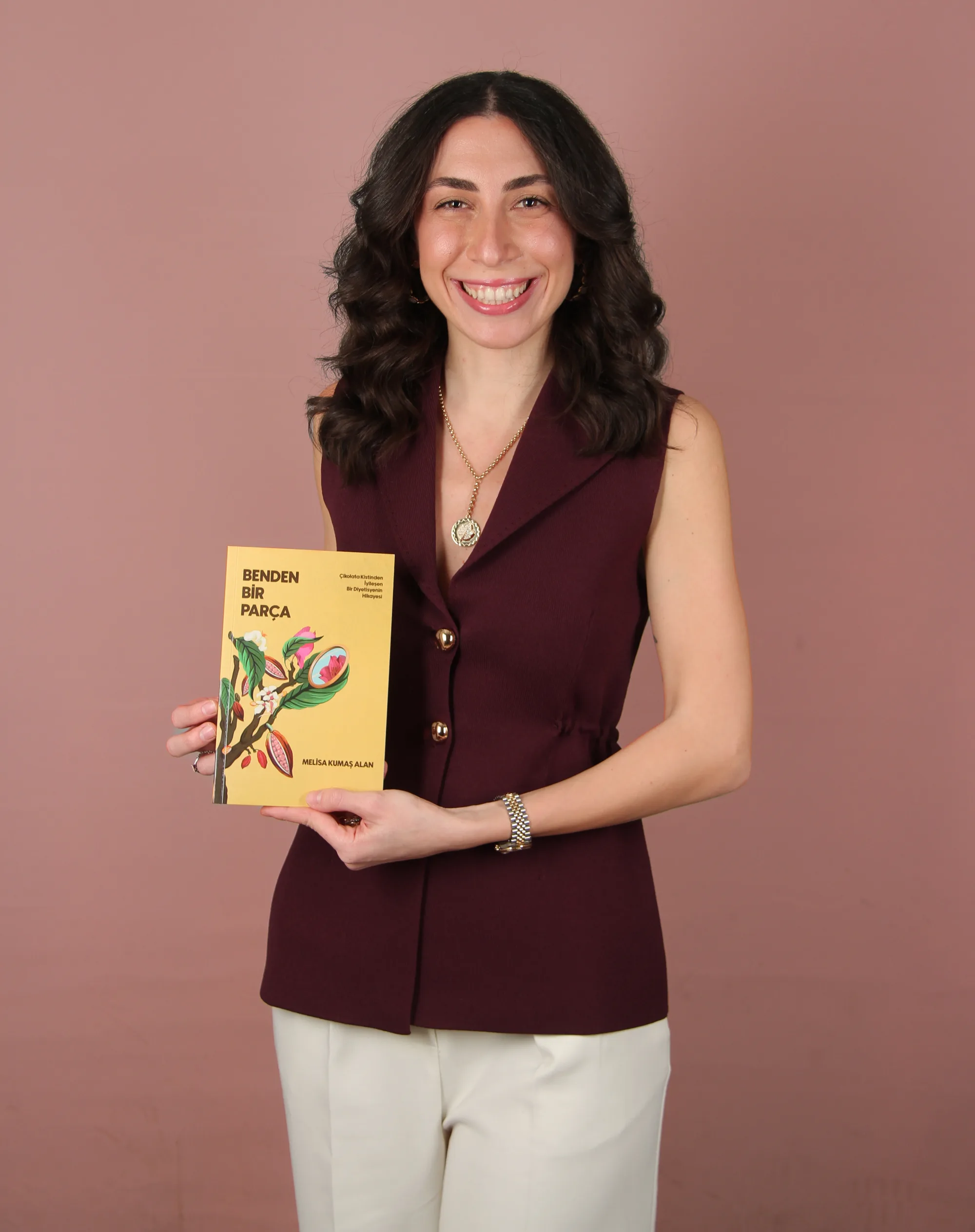 Dietitian Melisa Nadin Kumaş Alan smiling in front of a dusty pink backdrop, holding her book Part of Me