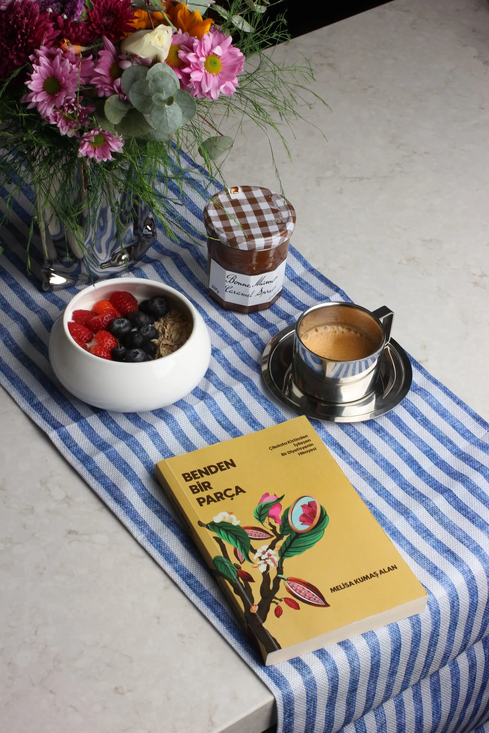 The book Part of Me on a blue striped table runner, beside a bouquet of flowers, a bowl of yoghurt with berries, a jar of caramel spread and a cup of coffee